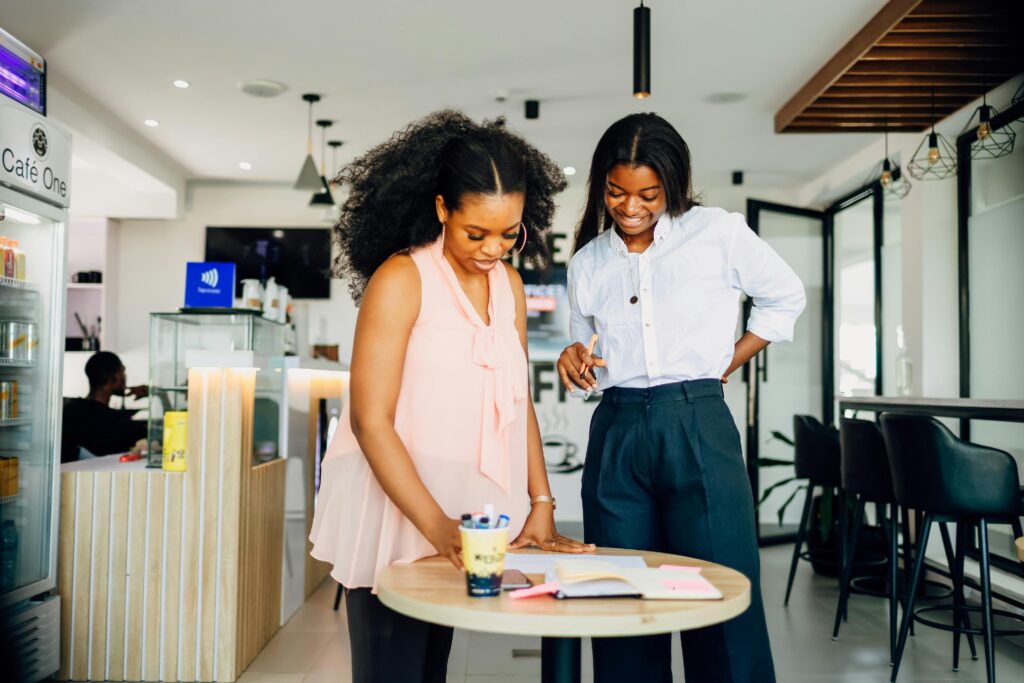 Two women stand next to a table in a cafe, working on a project while looking at a paper on the table. Find more afrocentric images on www.ninthgrid.com